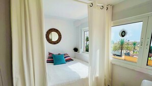Un dormitorio con cama, cortinas blancas, una ventana con vistas, un espejo redondo y una pequeña planta en maceta.