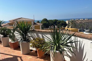 A rooftop terrace with potted plants, a view of the sea, and Mediterranean-style buildings.