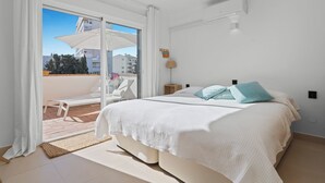 Un dormitorio con cama, mesita de noche, lámpara, puerta corrediza de vidrio que da a un balcón y vistas a edificios y árboles.