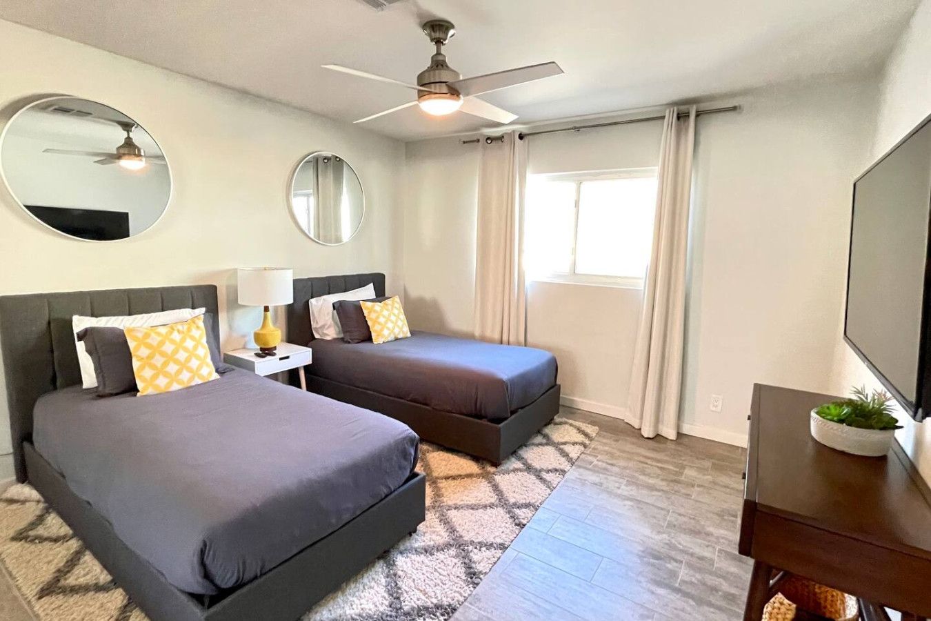 Quarto de hotel com duas camas, ventilador de teto, televisão e janela com cortinas.