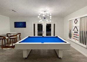 A pool table in a room with a modern chandelier, a pool table, and a rack of pool cues.