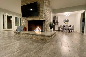 Ein modernes Wohnzimmer mit Kamin aus Stein, einem darüber hängenden Flachbildfernseher und einem Essbereich mit einer Deckenleuchte.