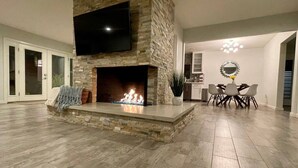 Uma sala de estar moderna com lareira de pedra, uma televisão de tela plana montada acima e um espaço de jantar com um candelabro.