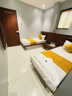 Une chambre d’hôtel avec deux lits, une tête de lit en bois, une table de chevet avec un téléphone et des bouteilles d’eau, et une fenêtre avec des stores.