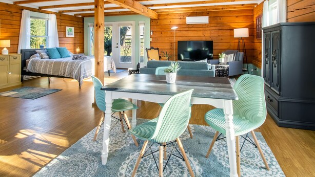 Ein gemütliches Blockhauszimmer mit Holzbalkendecke, einem Esstisch mit Stühlen, einer Couch und einem Fernseher.
