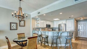 A kitchen with white cabinets, a central island with bar stools, a chandelier, and a dining area with wicker chairs.