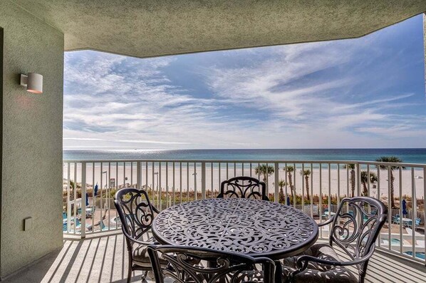 Un balcón con una mesa redonda y sillas, con vista a una playa con palmeras y cielo despejado.