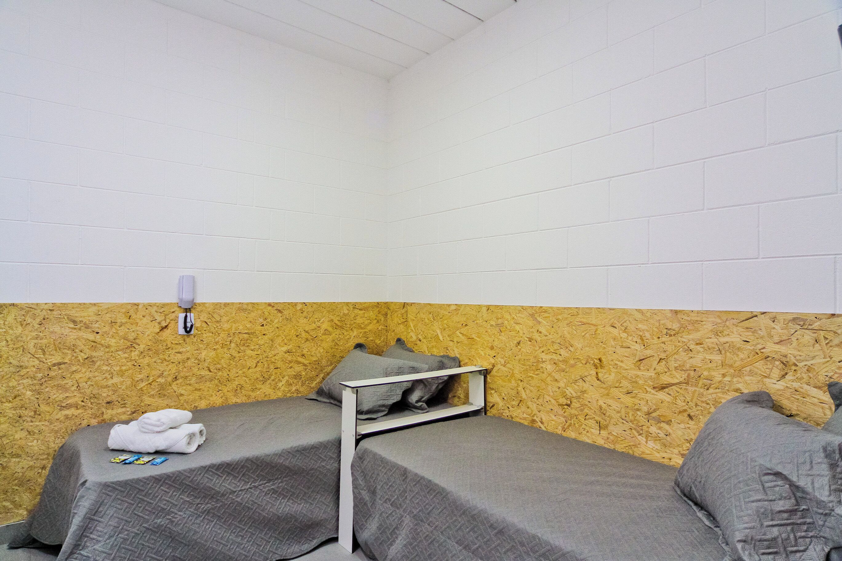 A small, single-room hotel room with two beds, a wooden wall, a white ceiling, and a wall-mounted light fixture.