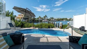 A pool area with lounge chairs, an umbrella, and a view of a body of water and buildings.