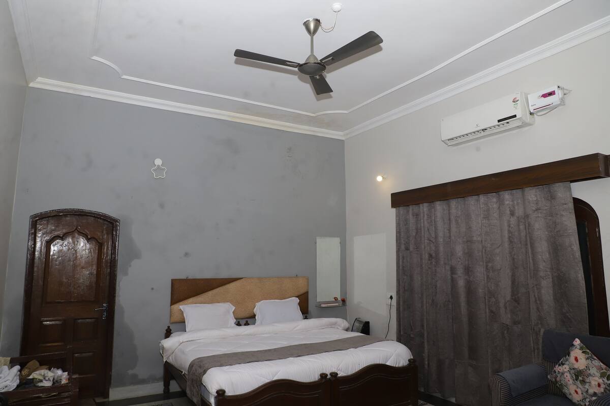 A bedroom with a bed, a ceiling fan, an air conditioning unit, a wooden wardrobe, and a curtain.