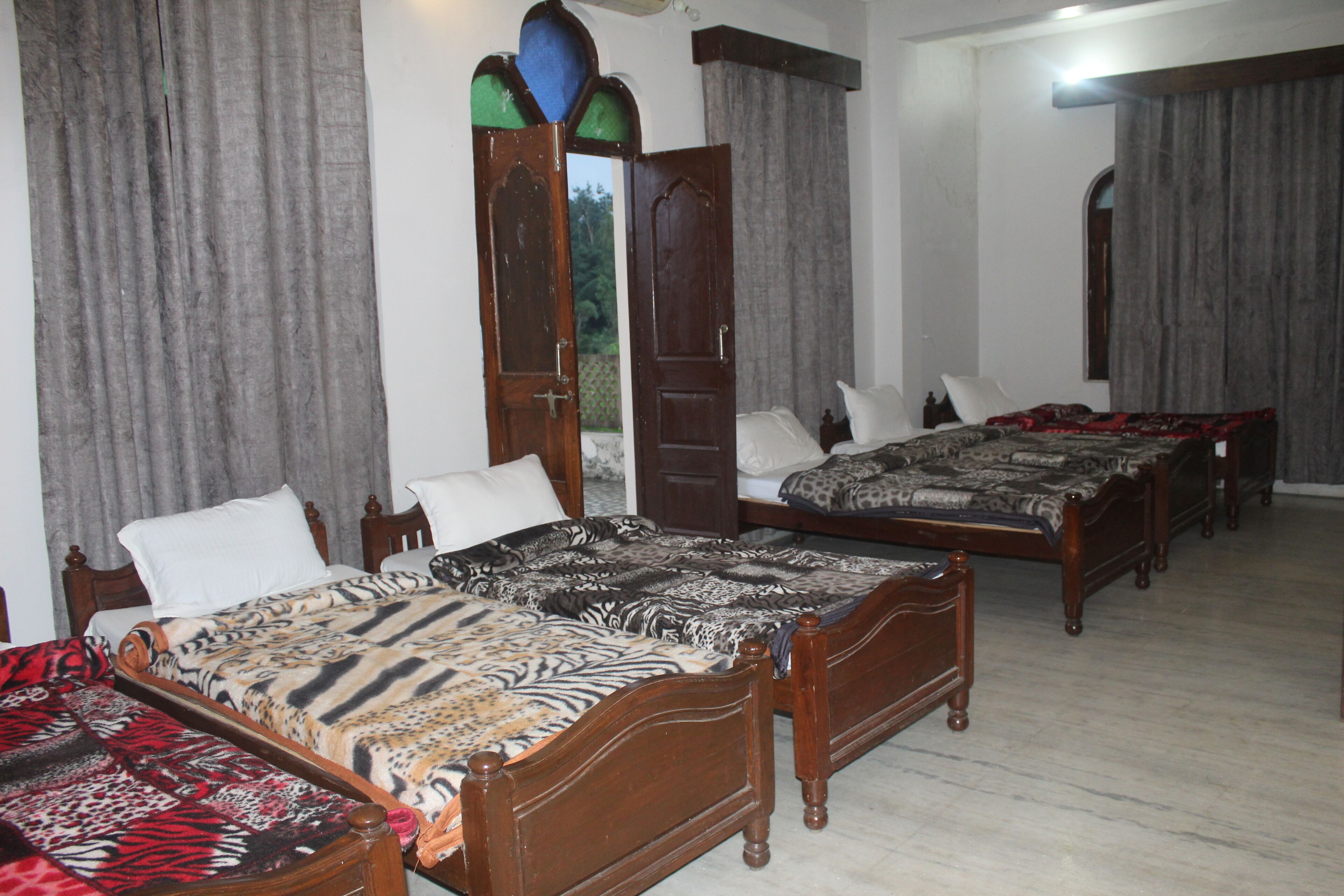 Quarto de hotel com três camas de madeira, cada uma com colcha estampada. O quarto tem piso de azulejos e janelas grandes com cortinas.