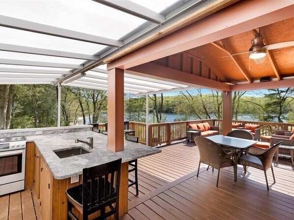 Um deck coberto ao ar livre com área de cozinha, assentos e vista para um lago.