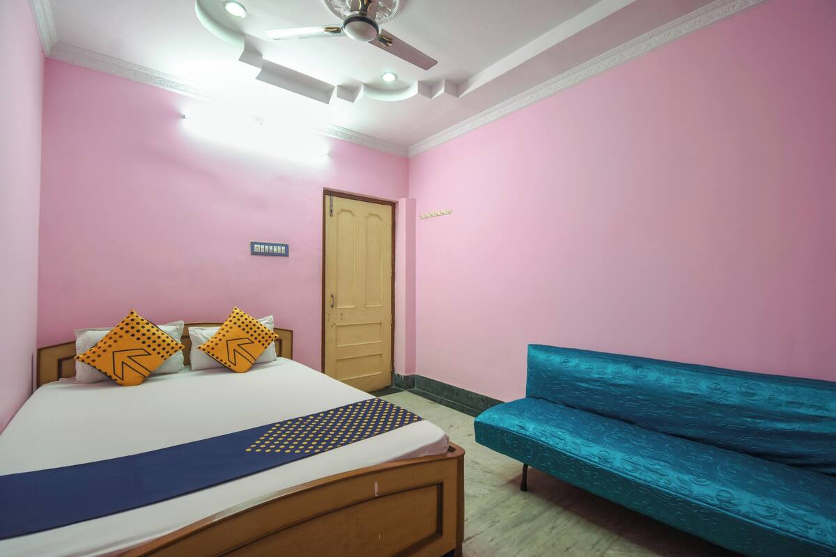 Quarto com paredes cor-de-rosa, cama de madeira, duas almofadas, colcha azul e um sofá azul, além de um ventilador de teto.