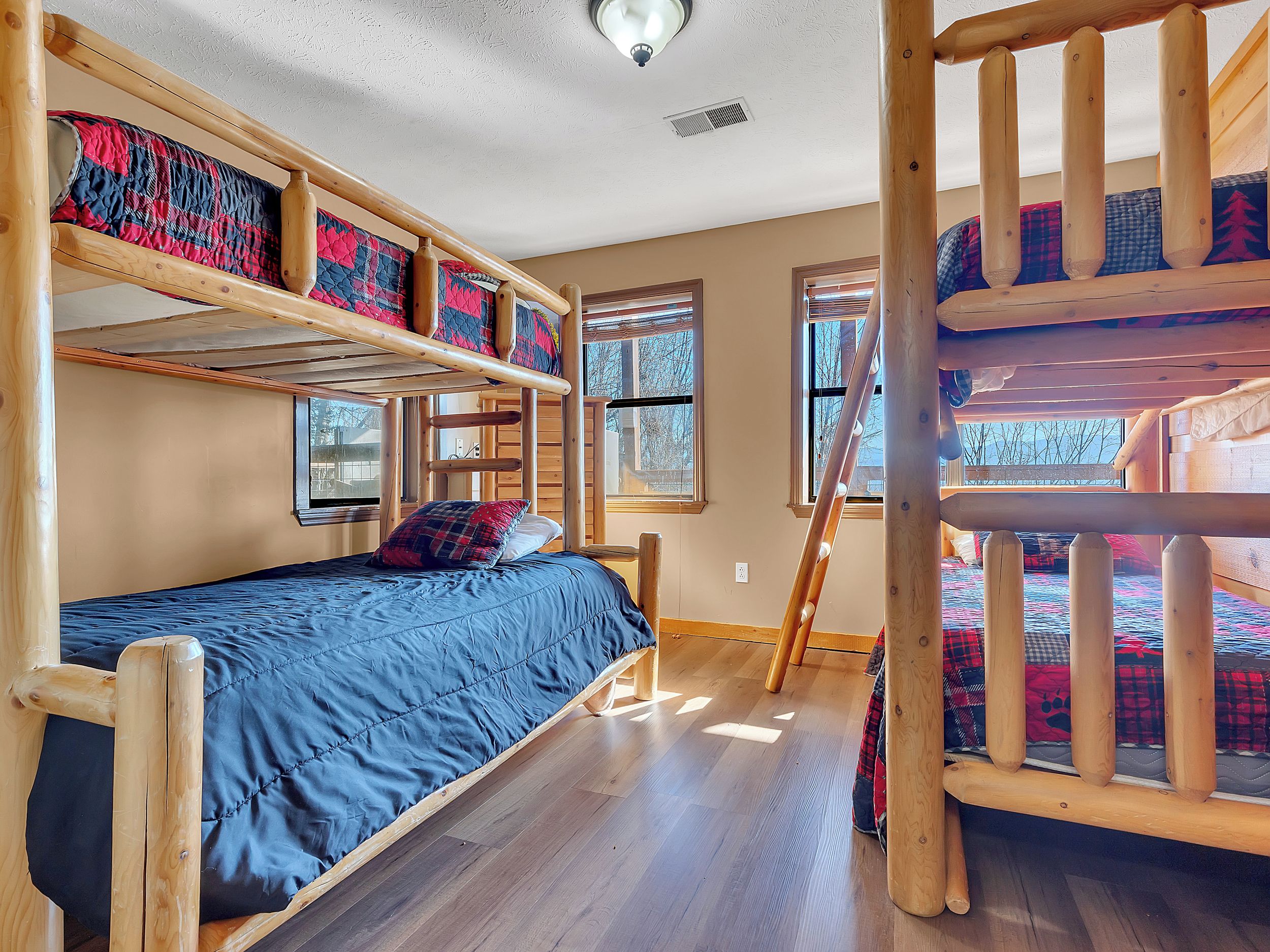 A bunk bed room with a blue bedspread, wooden railings, and windows with blinds.