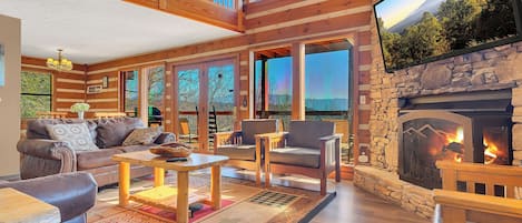 A spacious living room with a fireplace, wooden furniture, and large windows offering a view of the outdoors.