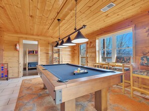 A pool table in a wooden room with a ceiling fan, a window with a view, and a door leading to another room.