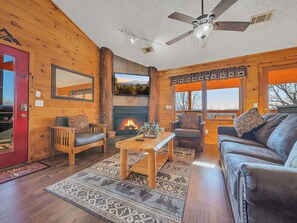 L’intérieur d’un chalet accueillant, doté d’une cheminée, de murs en bois, d’un ventilateur de plafond et d’une grande fenêtre offrant une vue panoramique.