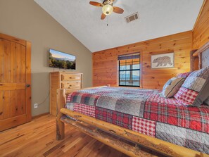Une chambre de style cabane en bois, avec un lit, une commode en bois, un ventilateur de plafond, une fenêtre et un téléviseur fixé au mur.