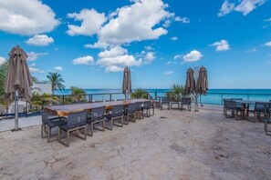 Eine Terrasse im Freien mit einem langen Holztisch und Stühlen, umgeben von Sonnenschirmen, mit Blick auf einen Strand.