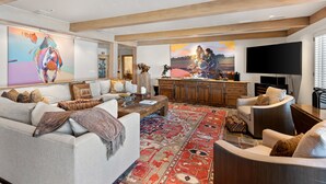 A spacious living room with a large sofa, a coffee table, and a TV. There is a wooden ceiling and a large rug on the floor.