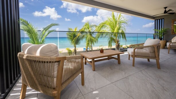 A balcony with wicker furniture, a wooden table, and a view of the beach and ocean. - Allure 202 2 Bedroom Apts (Bridgetown)