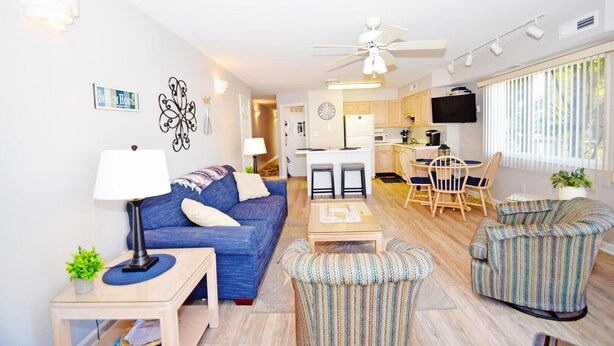 A modern living room with a blue sofa, a small table, and a patterned armchair. A ceiling fan and a TV are mounted on the wall. - Coral Ridge 1 2 Bedroom Condo (Ocean City)
