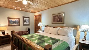 Un dormitorio con estructura de cama de madera, colcha verde y blanca, dos mesitas de noche con lámparas y cuadros enmarcados en las paredes.