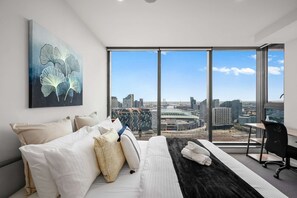 A modern hotel room with a large bed, a desk, and a view of a cityscape including a stadium. - Stadium Spacious Corner Unit with Perfect Location (Melbourne)