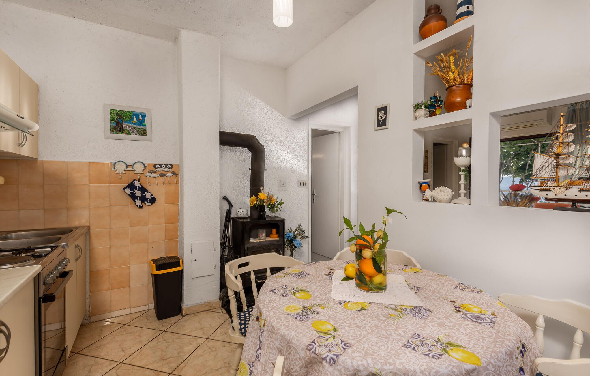Een keuken met een fornuis, oven en een betegelde spatwand. Er staat een eettafel met een bloemrijk tafelkleed en een bloevas.