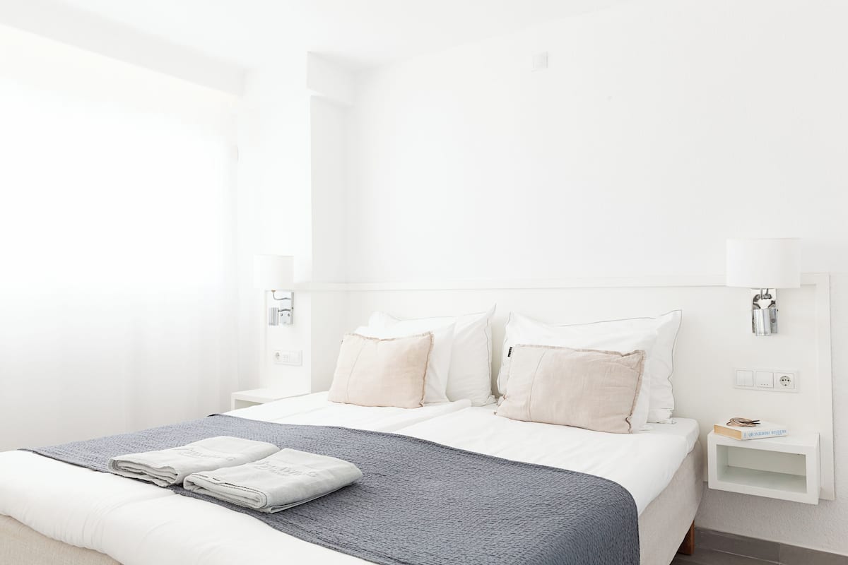 Un lit bien fait, avec une couverture grise et des oreillers, une table de chevet sur laquelle se trouvent un livre et une lampe, et un mur blanc avec un interrupteur.
