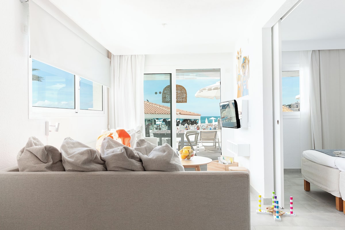 Une chambre d’hôtel moderne avec un canapé, une petite table sur laquelle sont disposés des fruits, une télévision et un balcon offrant une vue sur la ville.
