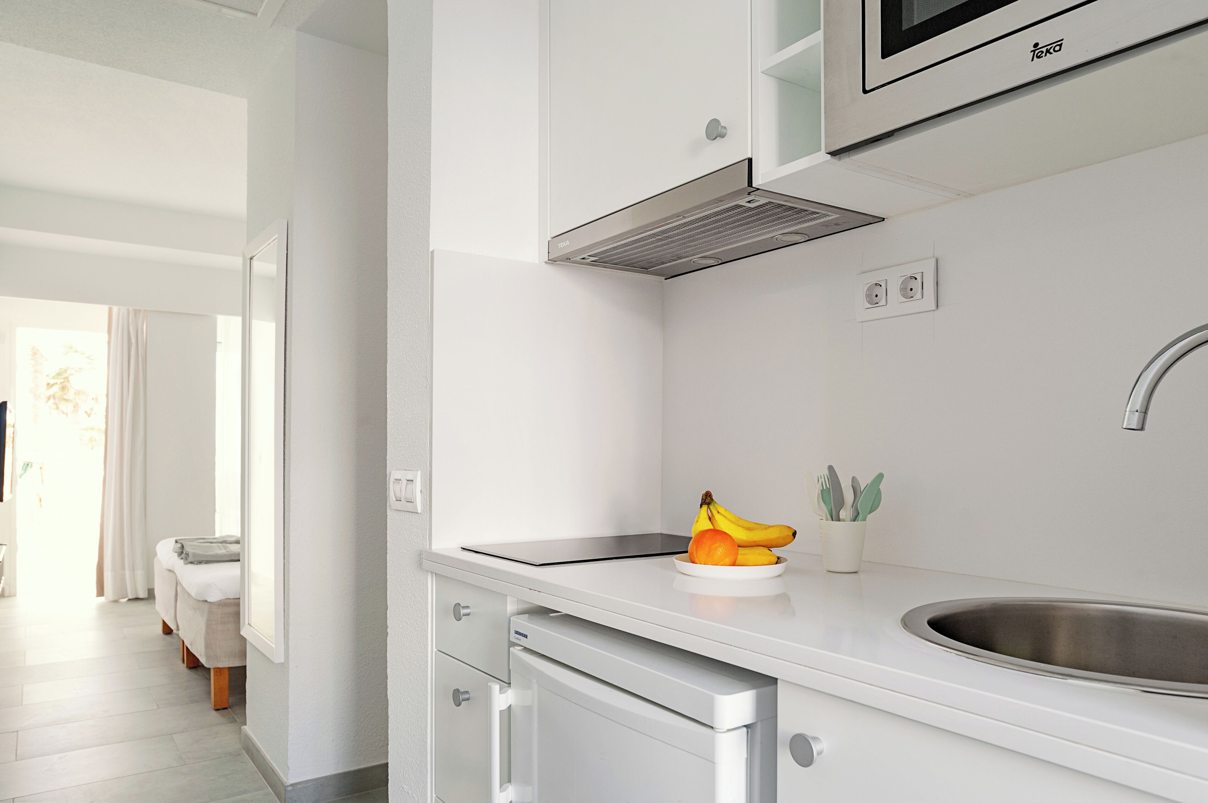 Cozinha moderna com armários brancos, micro-ondas embutido e pia de aço inoxidável. Há uma fruteira com bananas e um vaso de planta no balcão.