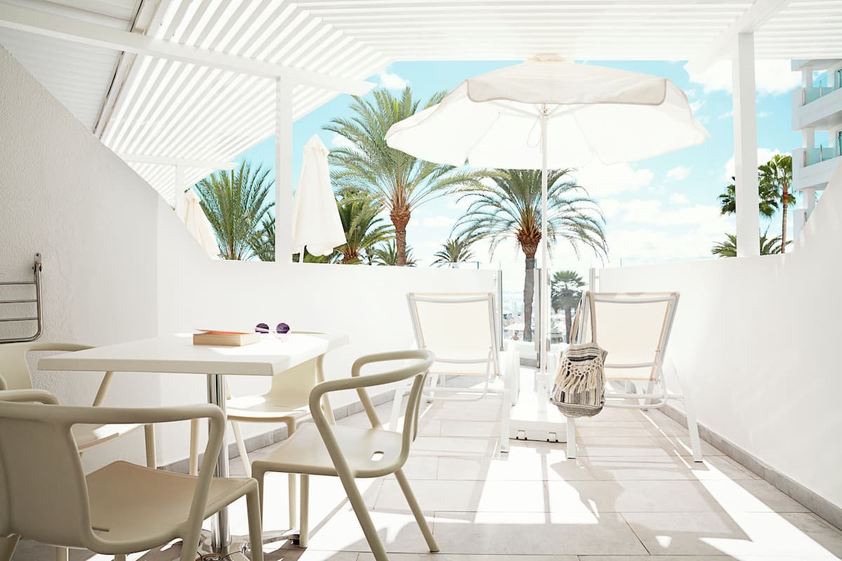 Un espace extérieur lumineux et ensoleillé, aménagé avec des tables et des chaises blanches, un grand parasol, et offrant une vue sur des palmiers et un bâtiment en arrière-plan.