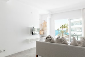 A modern living room with a large window, a flat-screen TV mounted on the wall, a sofa with cushions, and a small side table.