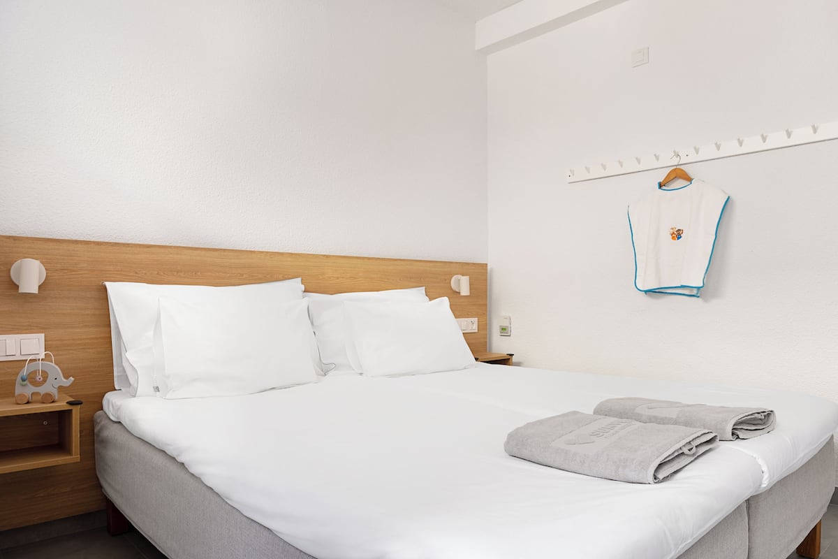 Un lit bien fait, avec du linge de lit blanc, une tête de lit en bois, une table de chevet avec une lampe et une serviette posée sur le lit.