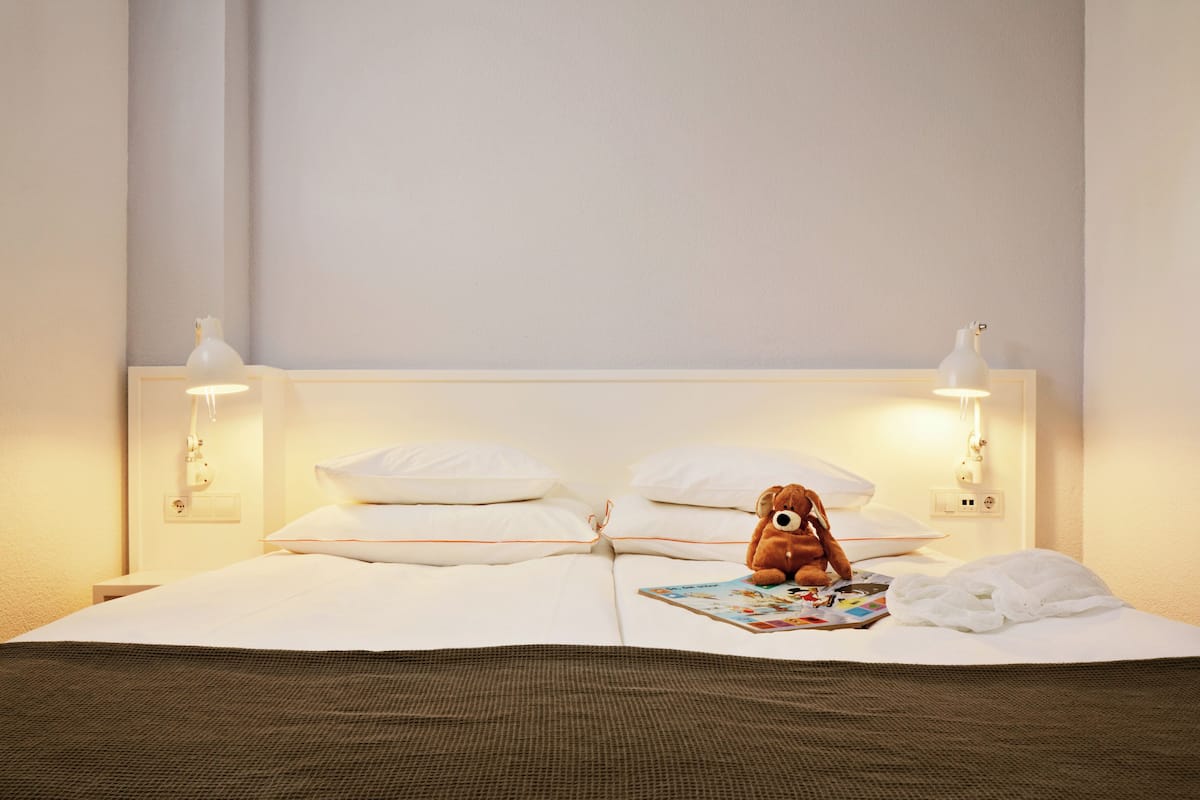 Un lit bien fait, avec des draps blancs, un ours en peluche marron et un magazine posé sur les oreillers.