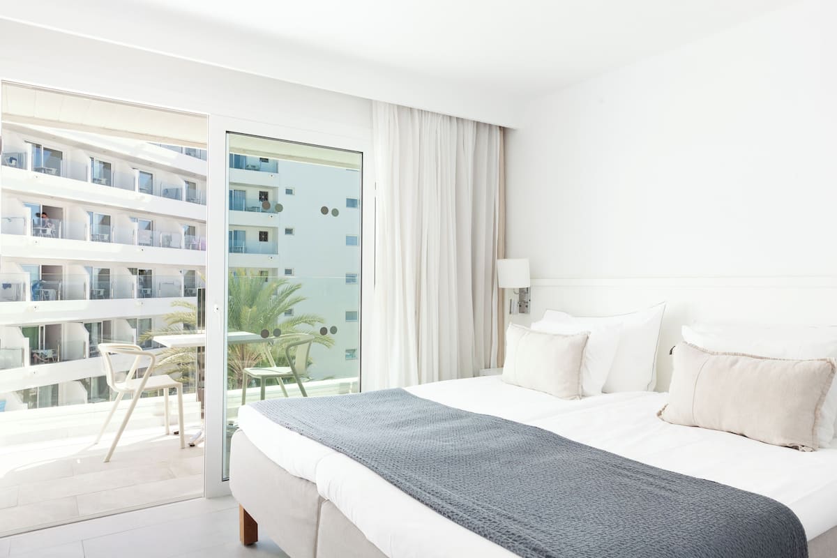 Une chambre d’hôtel avec un grand lit, un balcon doté d’une table et de chaises, et une vue sur d’autres immeubles.
