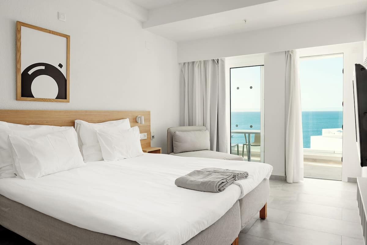 Une chambre d’hôtel avec un grand lit, une tête de lit en bois, une petite table avec une chaise et un balcon donnant sur la mer.