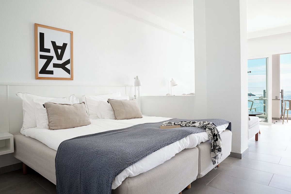 Une chambre moderne avec un grand lit, une vue sur la mer et une œuvre d’art encadrée au mur.