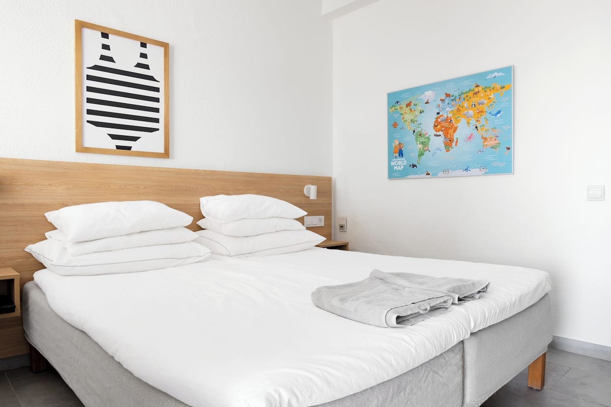 Un lit bien fait, avec du linge de lit blanc et des oreillers, une tête de lit en bois, une carte du monde accrochée au mur et une œuvre d’art abstraite encadrée.