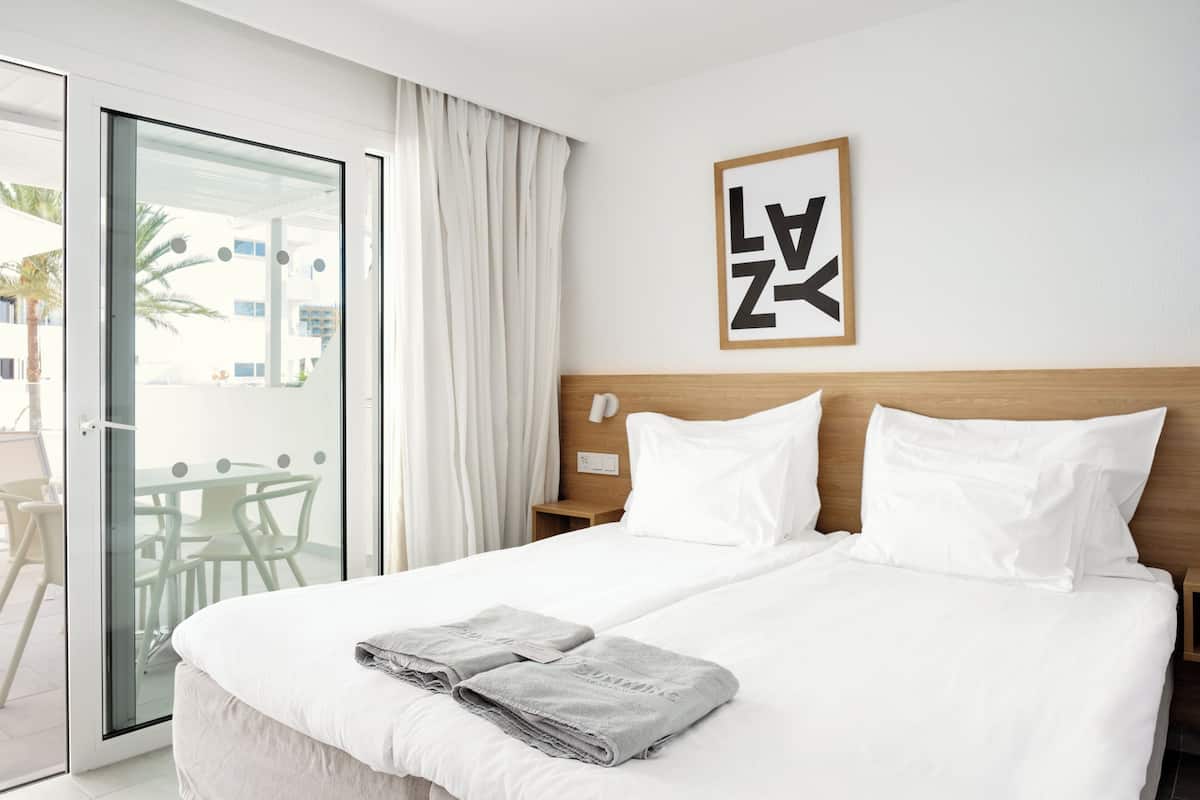 Une chambre d’hôtel avec un lit, du linge de lit blanc, une tête de lit en bois, une œuvre d’art encadrée et une porte-fenêtre coulissante donnant sur un espace extérieur avec une table et des chaises.