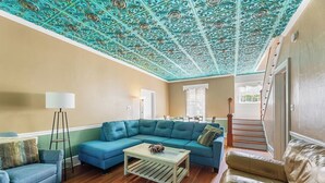 A living room with a teal-colored sectional sofa, a small coffee table, and a floor lamp. A staircase is visible in the background leading to another level.