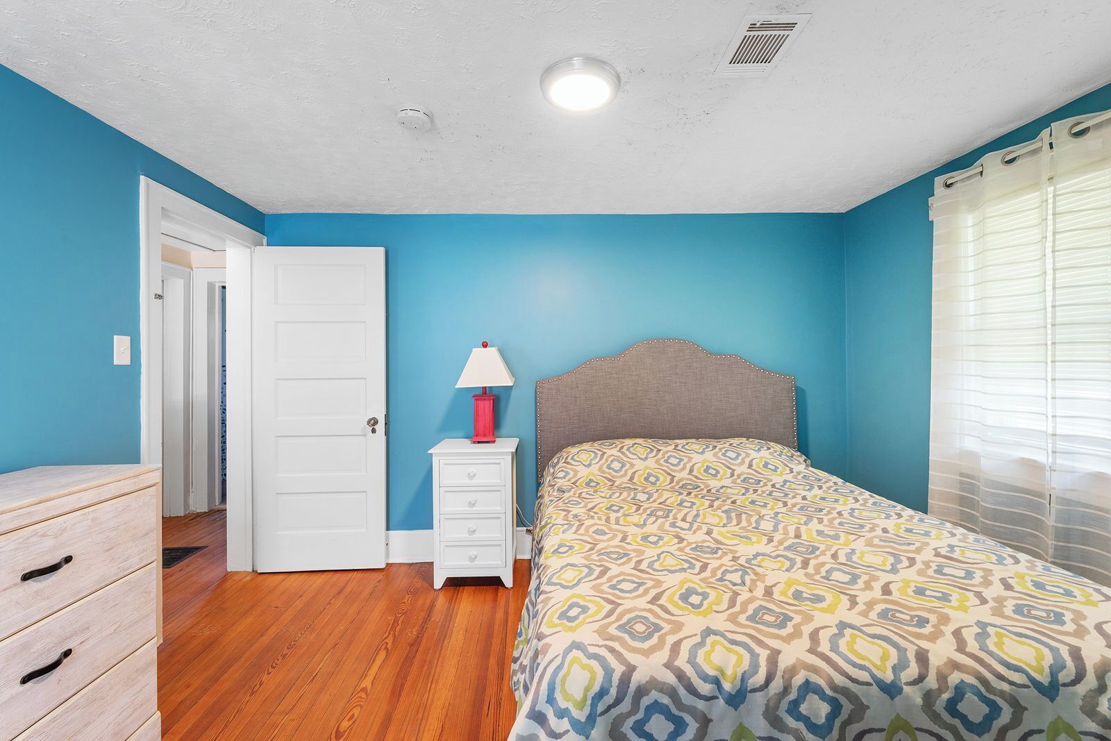 Een slaapkamer met een bed, nachtkastje, lamp, kledingkast en een deur.