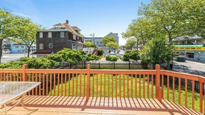A deck with a glass table, surrounded by a wooden fence and greenery, overlooking a street with buildings and a pizza shop.