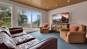 A spacious living room with a large window, leather sofas, and a flat-screen TV on a wooden cabinet. - The House - Waterfront Retreat: Secluded lodges perfect for nature lovers (Forge Creek)
