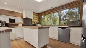 A modern kitchen with wooden flooring, white cabinetry, a central island, and a large window offering a view of trees. - The House - Waterfront Retreat: Secluded lodges perfect for nature lovers (Forge Creek)