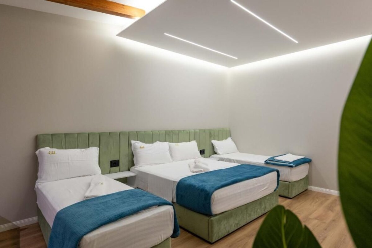 A hotel room with three single beds, each with a green headboard and white bedding. The room has a wooden ceiling and a large window.