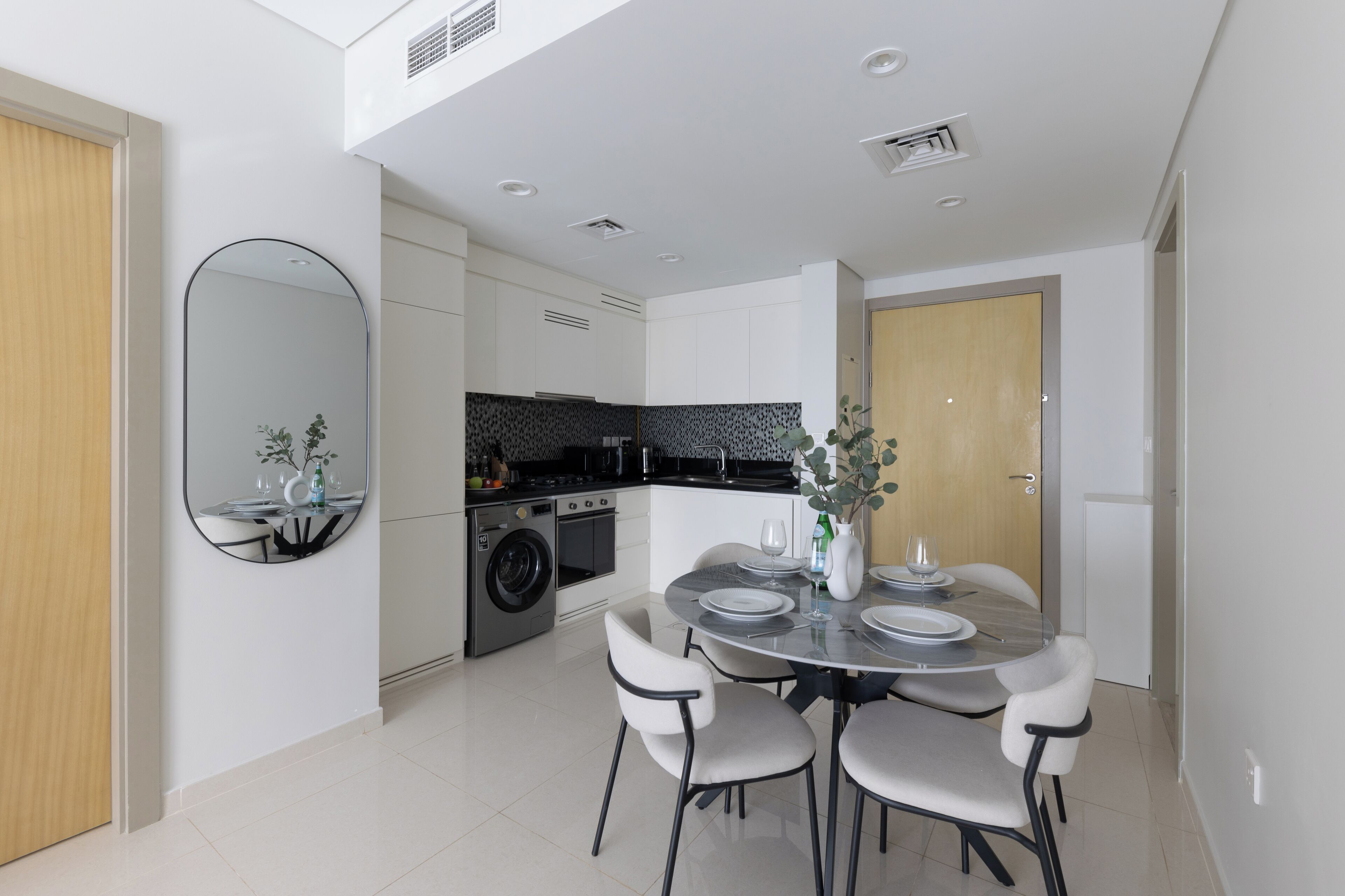A modern kitchen with a built-in washing machine, a dining area with a round table and chairs, and a large mirror on the wall.