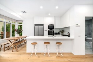 A modern kitchen with white cabinets, stainless steel appliances, and a wooden dining table with bar stools. - 3-Bed Luxury Apt in Port Melbourne (Port Melbourne)