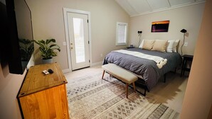 Un dormitorio con cama, banca, escritorio, planta en maceta y una puerta.